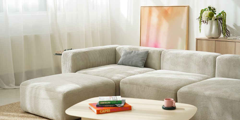 Beiges modulares Cordsofa in L-Form mit grauem Kissen, davor ein ovaler weißer Couchtisch mit Büchern und einer rosa Tasse, im Hintergrund helle Gardinen und ein abstraktes Wandbild in Rosa-Orange.